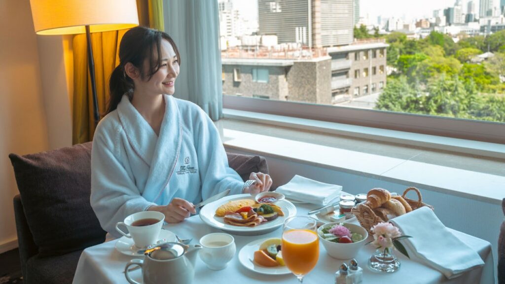 At the window table in the hotel room, a female guest is eating breakfast with a smile.