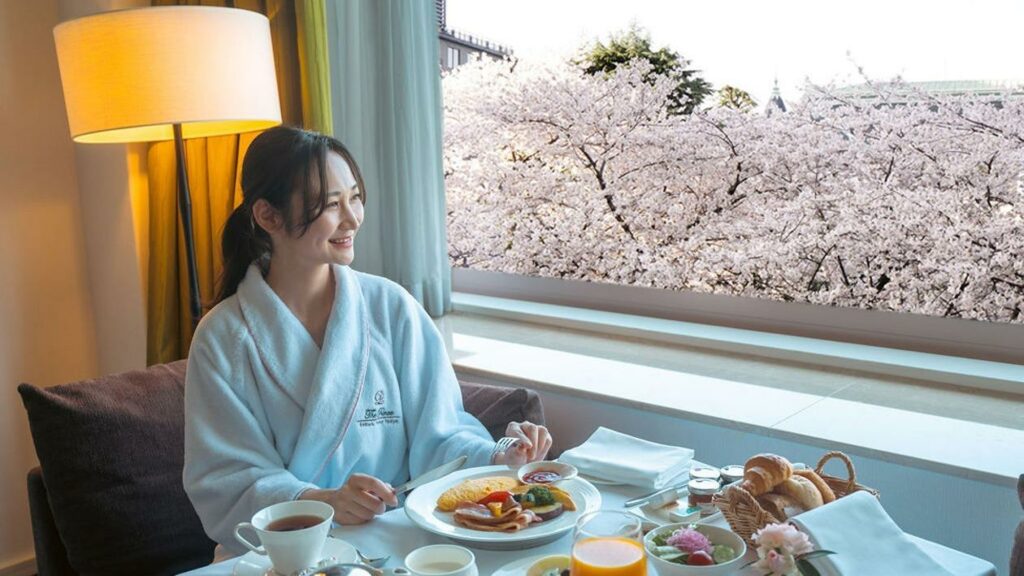 At the window table in the hotel room, a female guest is eating breakfast with a smile.