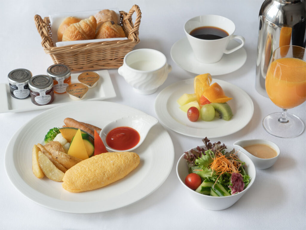 A Western-style breakfast is set on a white table, featuring an omelet, salad, fresh fruit, bread, coffee, and a glass of orange juice.